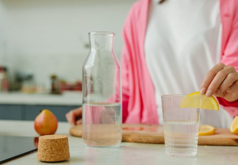 A woman in a pink outfit prepares lemon water in a bright, modern kitchen setting.